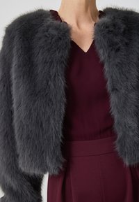 Mujer vistiendo una chaqueta de piel sintética gris oscuro sobre un vestido burdeos de escote en V con cintura alta y falda con plisados.