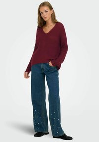 Maglione lavorato a maglia bordeaux con scollo a V, vestibilità ampia. Abbinato a jeans a gamba larga blu decorati con applicazioni di perle bianche all'orlo.