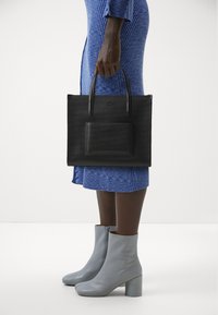 Black perforated tote bag with dual handles and a front pocket. Worn with a blue textured dress and gray heeled ankle boots.
