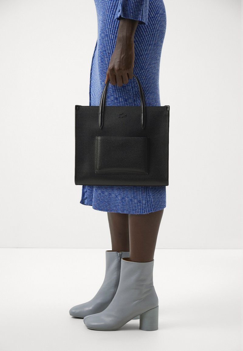 Black perforated tote bag with dual handles and a front pocket. Worn with a blue textured dress and gray heeled ankle boots.