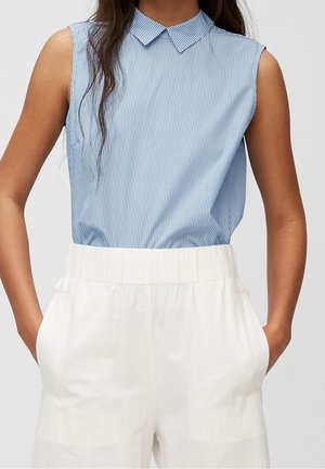 Mujer vestida con un top sin mangas de rayas azules y blancas con cuello, metido en pantalones blancos de talle alto, con las manos en los bolsillos.