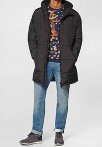 Schwarzer, langer, gepolsterter Mantel mit Kapuze, getragen über einem farbenfrohen, gemusterten Pullover, kombiniert mit hellblauen Jeans und grauen Turnschuhen.