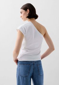 Weißes, asymmetrisches T-Shirt mit kurzen Ärmeln, aus Baumwolle. Kombiniert mit hellblauen Jeans mit klassischem Gesäßtaschendesign.