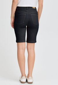 Pantaloni Bermuda in denim nero con vita alta, tasche posteriori e orlo arrotolato, abbinati a sneakers bianche su uno sfondo chiaro.