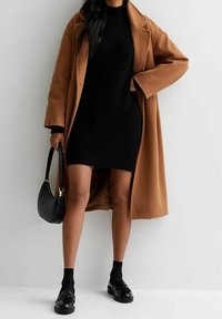 Cappotto marrone su un vestito in maglia nera, abbinato a calze nere e mocassini neri lucidi; include una piccola borsa nera.
