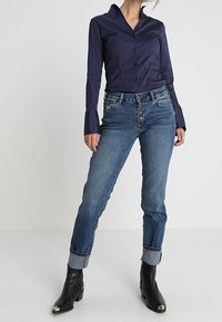 Granatowa koszula zapinana na guziki z wywijanym kołnierzem, zestawiona z niebieskimi dżinsami z pięcioguzikowym zapięciem i podwiniętymi mankietami. Czarne botki za kostkę z metalowymi czubkami.