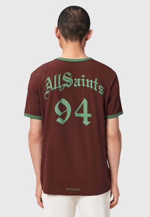 Mand iført brun kortærmet skjorte med grøn kant, teksten "AllSaints", nummeret "94" og romertal "MCMXCIV" trykt på ryggen.