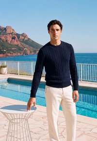 Homme en pull marine et pantalon blanc se tient au bord de la piscine en tenant une petite table ronde blanche avec l’océan et des collines rocheuses en arrière-plan.