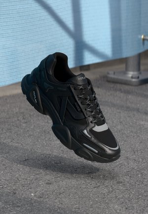 Zwarte en grijze REPLAY sneaker met reflecterende details, zwevend boven asfalt nabij een lichtblauwe textuurmuur.