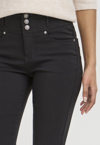 Jeans de mezclilla negra con una cintura alta, cierre de tres botones y herrajes plateados. Presenta bolsillos laterales y una textura suave y elástica.