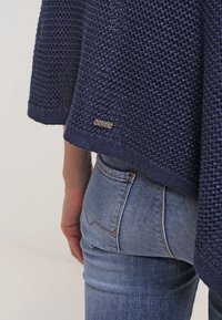 Marinblå stickad tröja med ett strukturerat mönster, med en etikett på nederkanten. Kombinerad med ljusblå denimjeans.