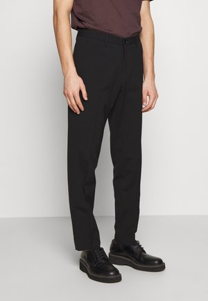 Uomo che indossa pantaloni neri su misura e scarpe nere con lacci, in piedi contro uno sfondo chiaro e uniforme.