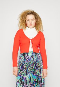 Cardigã vermelho com nervuras, com um fecho atado à frente, usado sobre um suéter de gola alta branco, combinado com calças largas de padrão multicolorido.