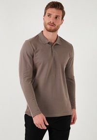Camisa tipo polo de manga larga, color marrón, con cuello y dos botones, confeccionada en una tela texturizada, combinada con pantalones oscuros.