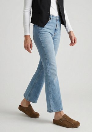 Person wearing light blue denim jeans, brown slip-on clogs, white long-sleeve top, and black buttoned vest walking on a plain background.