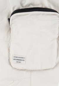 Vitt tyg med en ficka med dragkedja som har svarta detaljer. En etikett med texten "Levi Strauss & Co. Est. 1873" står ovanför geografiska koordinater.