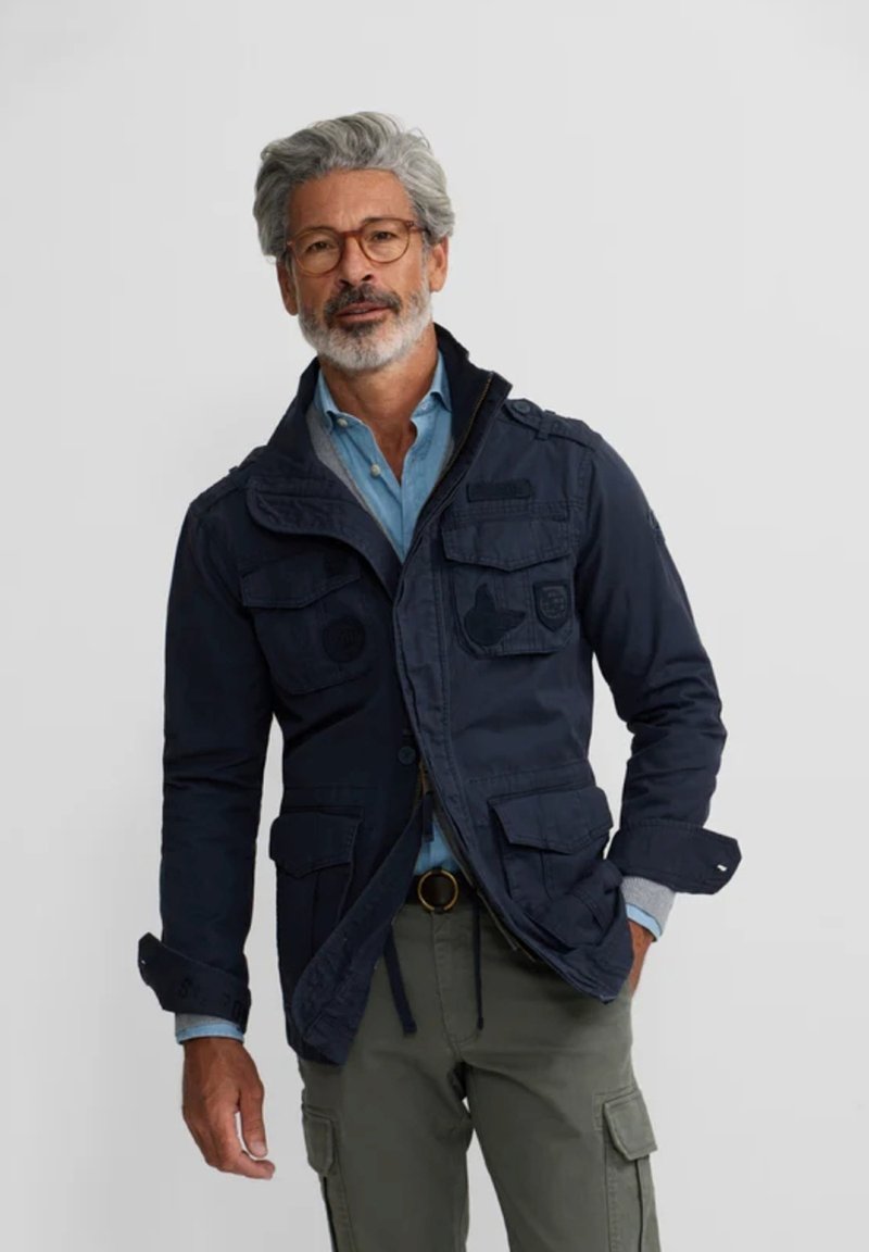 Chaqueta de marina hecha de tela duradera con múltiples bolsillos, un cierre frontal con cremallera y puños ajustables. Llevada sobre una camisa de color azul claro.