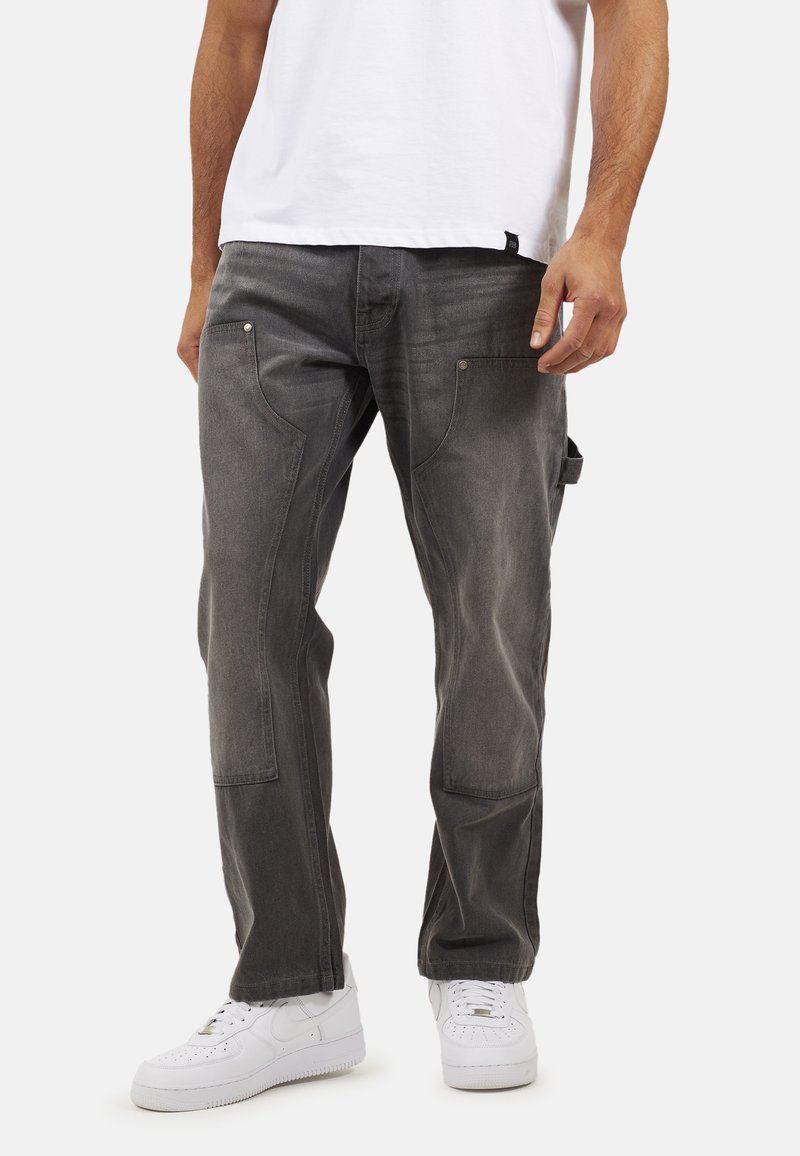 Homme portant un pantalon de charpentier gris foncé et des baskets blanches, se tenant contre un fond blanc avec une main détendue le long du corps.