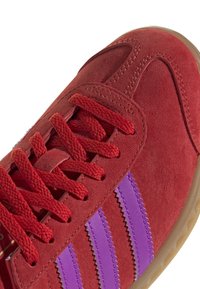 Czerwony zamszowy sneaker z fioletowymi paskami, teksturowaną powierzchnią, zamkniętym systemem sznurowania i gumową podeszwą. Posiada szwy w tonacji oraz wzmocniony nosek.