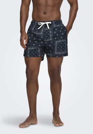 Torso y piernas masculinas con pantalones cortos de baño azul marino con estampado blanco de bandana paisley y cordón blanco, de pie descalzo sobre un fondo liso.
