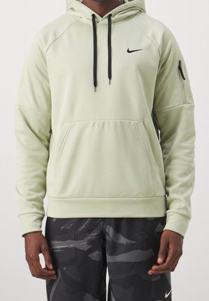 Person iført en lysegrøn Nike hættetrøje med sorte snore og sorte Nike camouflage-shorts, stående mod en ensfarvet baggrund.