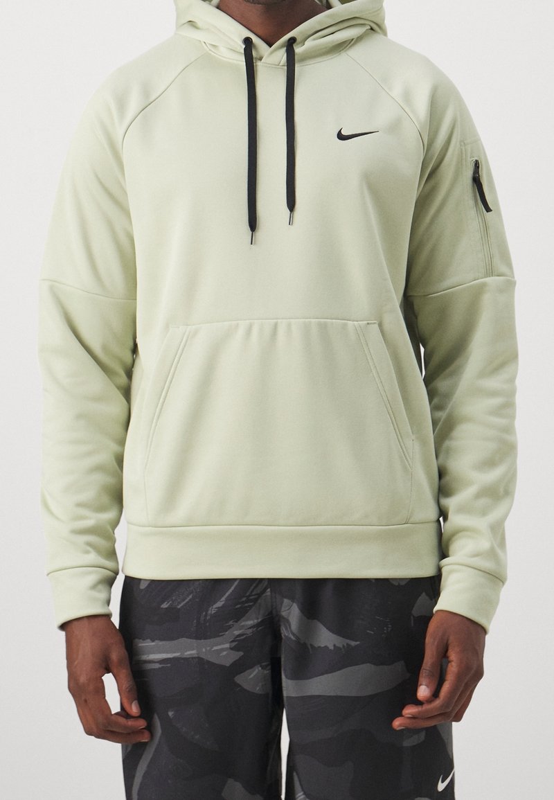 Personne portant un sweat à capuche Nike vert clair avec des cordons noirs et un short Nike camouflage noir, debout devant un fond uni.