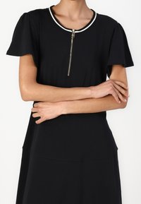 Musta mekko lyhyillä hihoilla, jossa on valkoisella viimistellyt pyöreä pääntie ja edessä vetoketju. Pehmeä, sileä kangas ja laskostava malli.