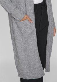 Cardigan en tricot gris avec des poignets côtelés et un design à ouverture devant, doté d'une poche latérale et porté par-dessus un pantalon noir.