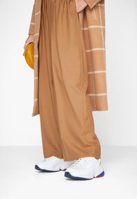 Vita sneakers med blåa och röda accenter, i kombination med lösa beige byxor och en lång kamelhudskappa med vita ränder, hållande en gul väska.