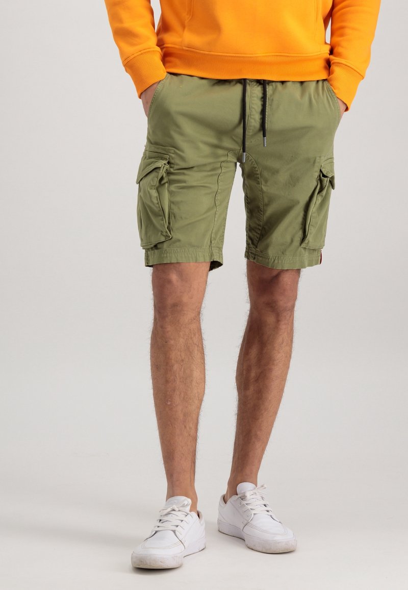 Homme portant un short cargo vert olive, des baskets blanches et un haut à manches longues orange, les mains dans les poches, debout sur un fond uni.