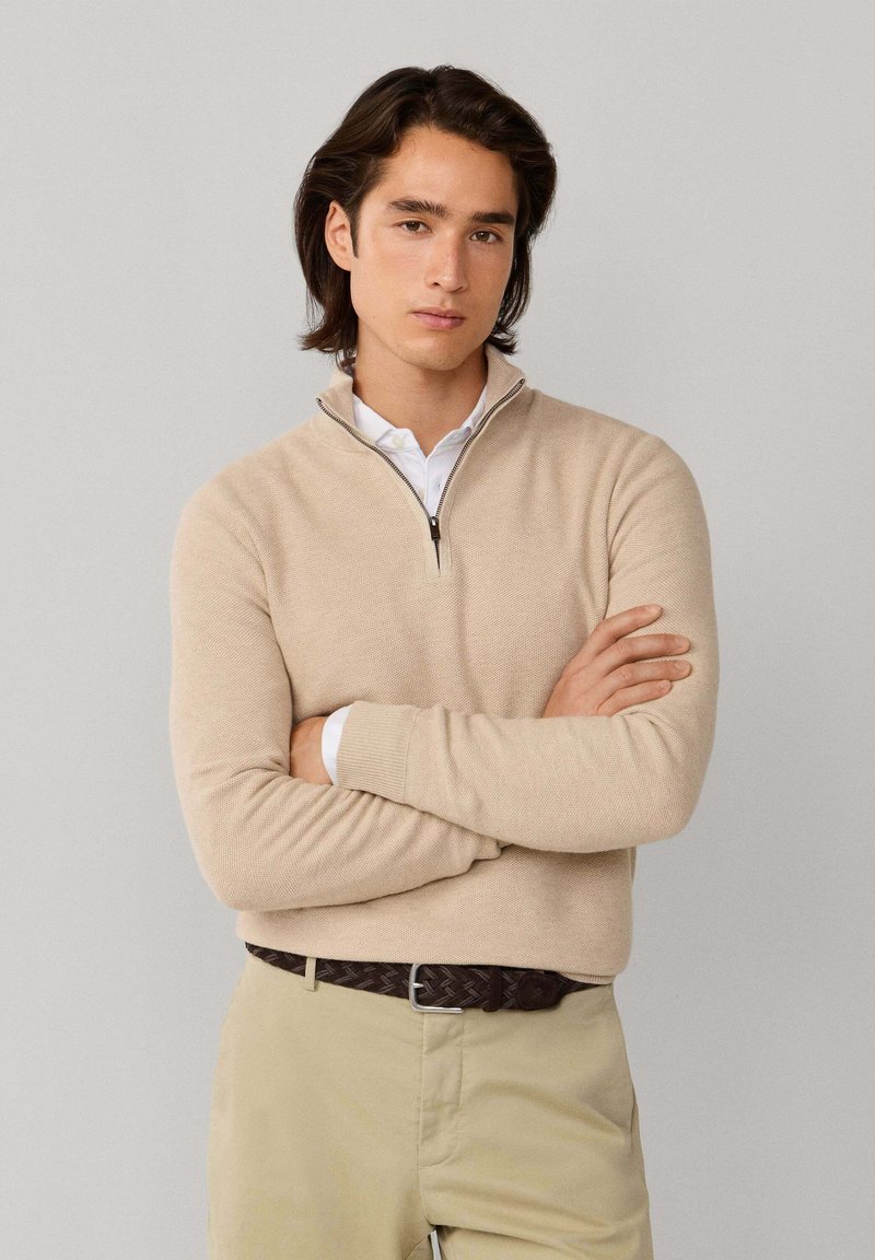 Mladý muž so stredne dlhými tmavými vlasmi má na sebe béžový sveter na zips cez bielu košeľu, skrížené ruky a svetlé nohavice so svetlohnedým opaskom.