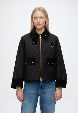 Femme portant une veste matelassée noire avec une fermeture éclair et des boutons dorés, debout devant un fond clair uni, associée à un jean bleu.