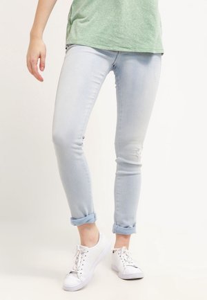 Personne portant un jean skinny bleu clair retroussé, des baskets blanches et une chemise verte à manches courtes, debout avec une jambe légèrement avancée.