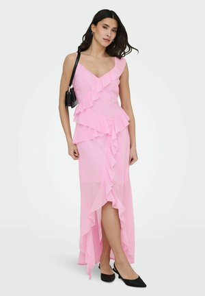 Kobieta stojąca w różowej, bezrękawnikowej sukience z falbanami i asymetrycznym dołem, czarne buty typu slingback oraz nosi małą czarną torebkę na ramię.