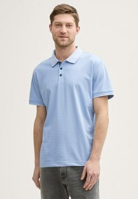 Ljusblå polo med krage och knappslå med tre knappar; kort ärm; slät textur; matchad med grå jeans.
