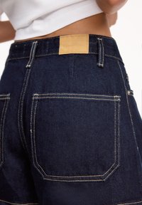 Pantalones cortos de mezclilla azul oscuro con cintura alta, con costuras amarillas en contraste, un parche de logo de cuero y dos bolsillos traseros.