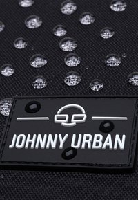 Fekete szövet táska vízcseppekkel a felszínen. Négyzet alakú címke található rajta, amelyen a "JOHNNY URBAN" felirat fehér színnel, fekete háttérrel van írva.