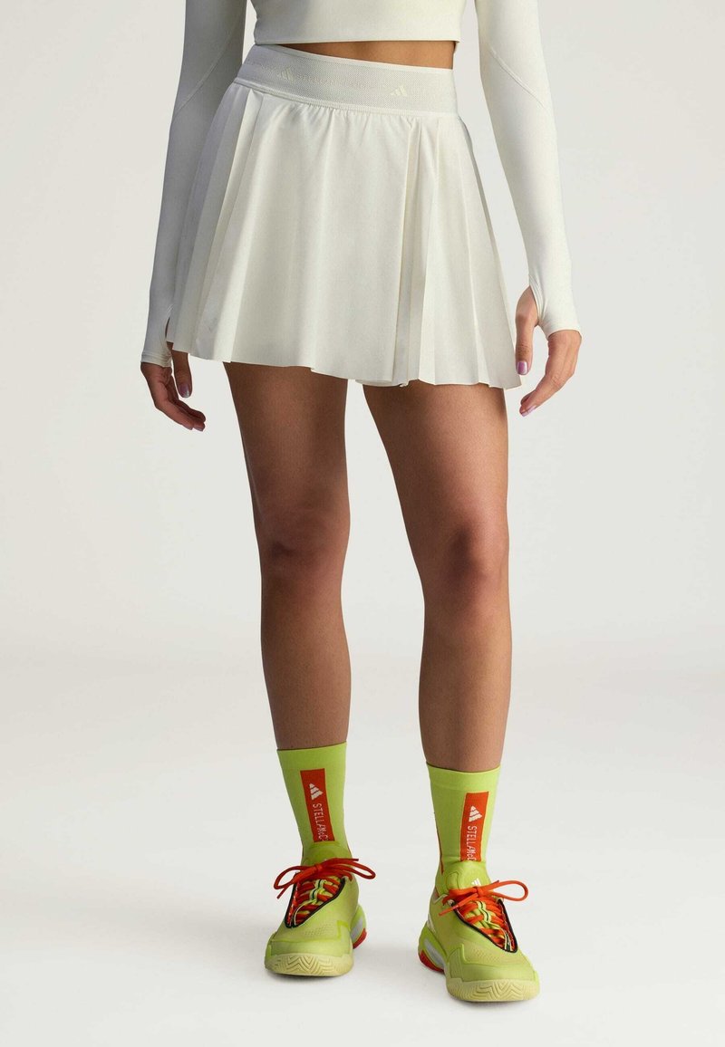 White pleated skirt with a wide waistband, paired with bright green socks and sneakers featuring red-orange accents and textured details.