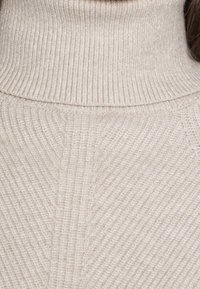 Maglione beige a collo alto a costine con un alto colletto, texture morbida e un sottile motivo diagonale nel tessuto.