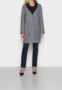 Donna che indossa un lungo cappotto a quadri bianco e nero, jeans scuri e décolleté neri a punta, in piedi contro uno sfondo bianco semplice.