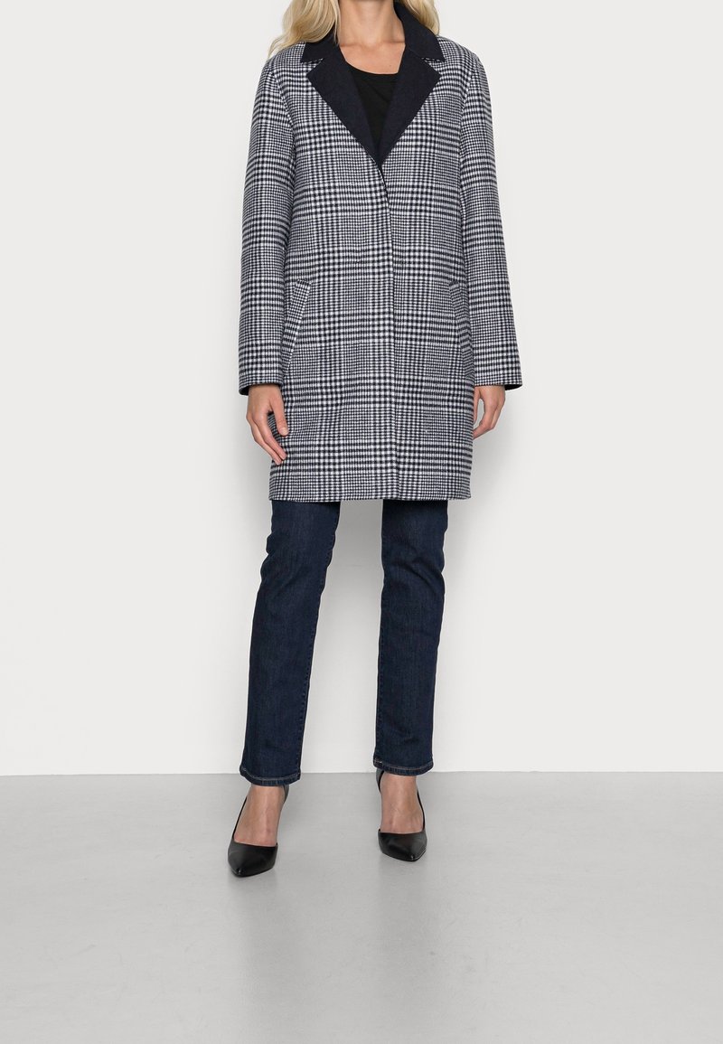 Donna che indossa un lungo cappotto a quadri bianco e nero, jeans scuri e décolleté neri a punta, in piedi contro uno sfondo bianco semplice.