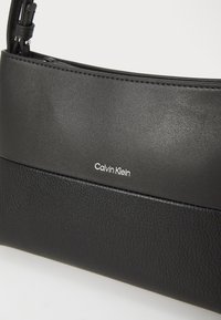 Czarna torebka na ramię z syntetycznej skóry o gładkiej fakturze, z kontrastowym panelem i metalowym logo "Calvin Klein". Zamek błyskawiczny.