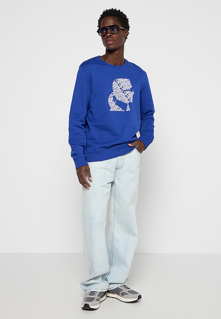 Sweatshirt bleu royal avec un design graphique blanc sur le devant, associé à un jean loose bleu clair et des baskets grises. Tissu texturé, coupe décontractée.