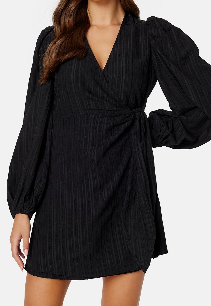 Vestido negro envolvente con escote en V, mangas largas abullonadas, rayas verticales texturizadas y un detalle de lazo lateral que acentúa la cintura.