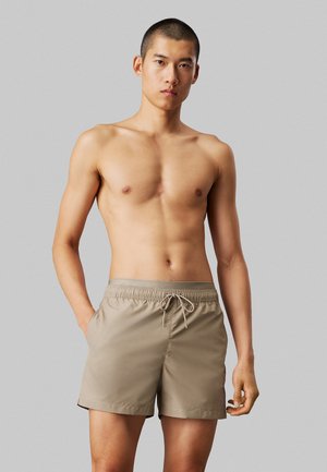 Tan badetights med elastisk livrem og snor, der har sidelommer og en glat stofstruktur. Modellen har kort hår.