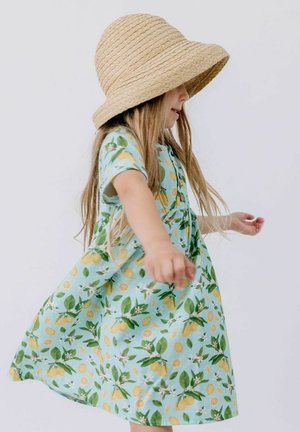 Junges Mädchen, das sich in einem hellblauen Kleid mit Zitronen- und Blätterdruck dreht, trägt einen breitkrempigen, geflochtenen Strohhut vor einfachem Hintergrund.