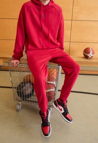 Hoodie rouge et pantalon de survêtement avec un logo Jordan, assortis à des baskets montantes noires et rouges, assis dans un chariot de supermarché avec des ballons de basket.