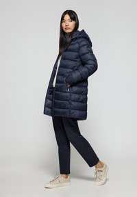 Marinblå vadderad jacka med huva, med dragkedjestängning och framfickor, bärs över en ljusgrå tröja och mörka byxor.