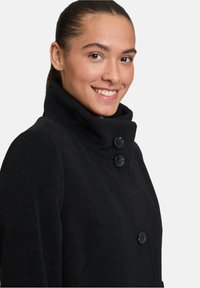 Cappotto di lana nera con colletto alto, dotato di due grandi bottoni neri, tessuto strutturato e un design aderente che si restringe in vita.
