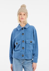Veste en polaire bleue avec col, fermeture à boutons sur le devant, deux poches plaquées et tissu texturé. Assortie à un bonnet en tricot beige.